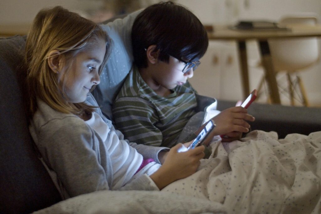 2 enfants sont leur lit avec un écran dans les mains